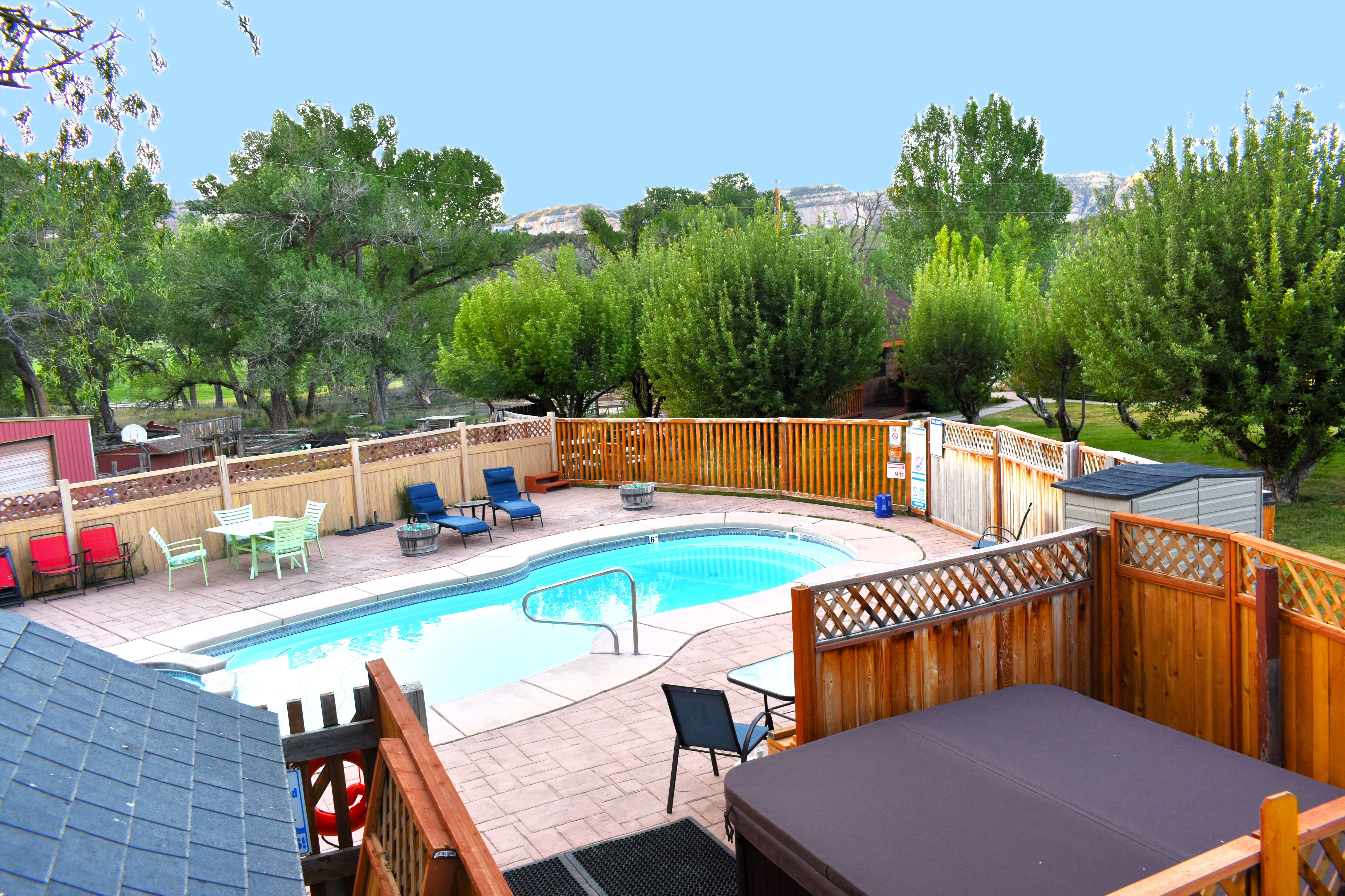 A fenced pool area with a pool, hot tub, and outdoor furniture surrounded by trees and a mountain view.